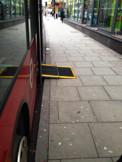 A red London bus at a stop with its wheelchair access ramp folded out onto the pavement