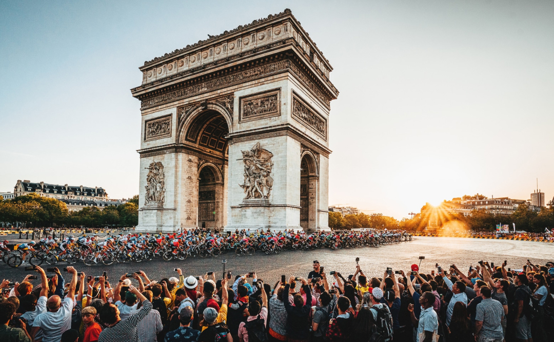 Tour de France peloton passing the Arc de Triomphe at sunset as crowds photograph them.