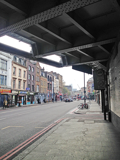 London street running under a steel railway bridge, lined with shops and a 20 mph zone sign