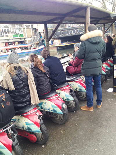 People sitting on a row of scooter seats beside a canal at Camden, houseboats and a bridge behind