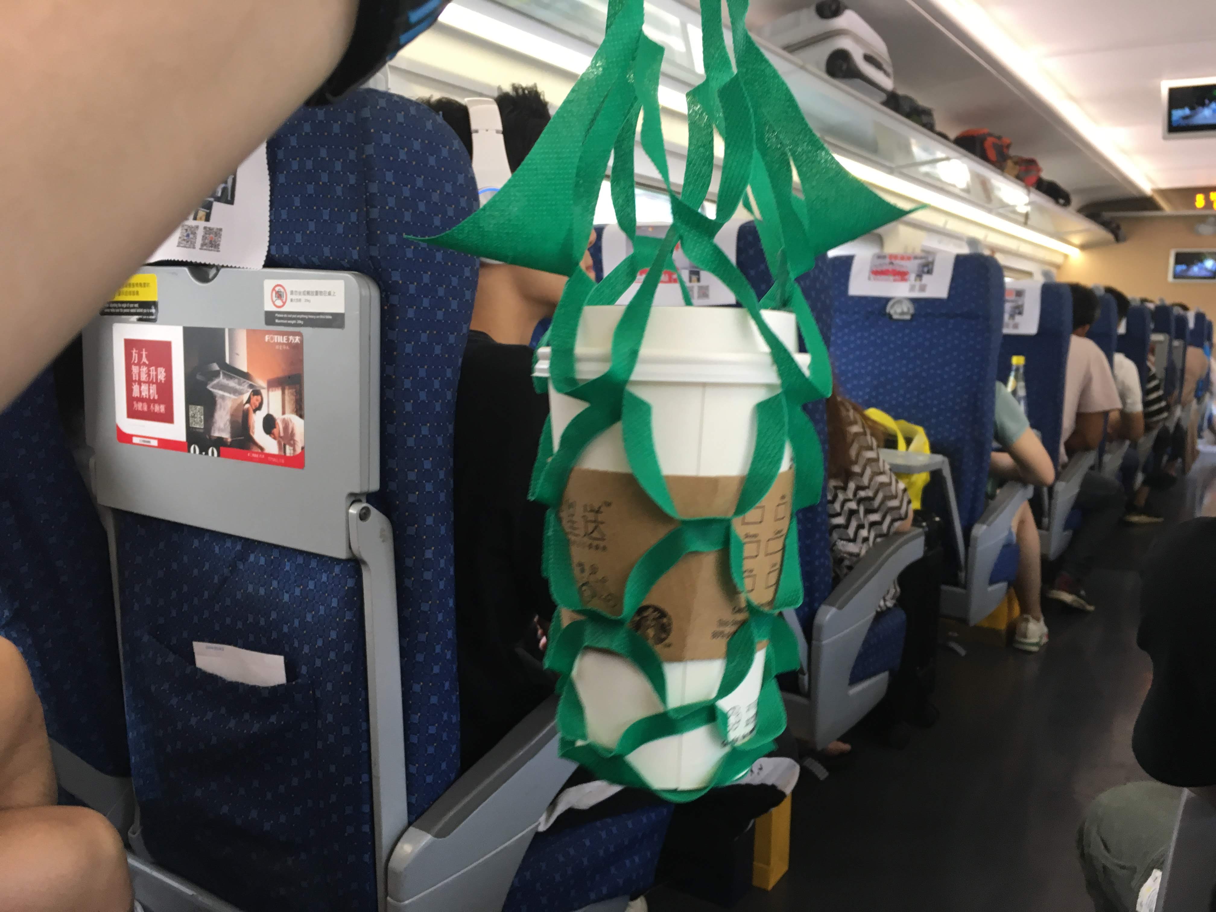 Two takeaway coffee cups carried in a green mesh bag hanging in a crowded train aisle.